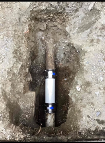 A buried metal pipe joint exposed in a dirt trench, with a white cylindrical coupling in the center.