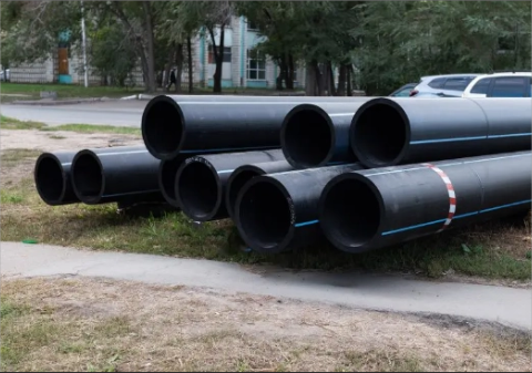 Stacked large black pipes beside a road with trees and buildings in the background