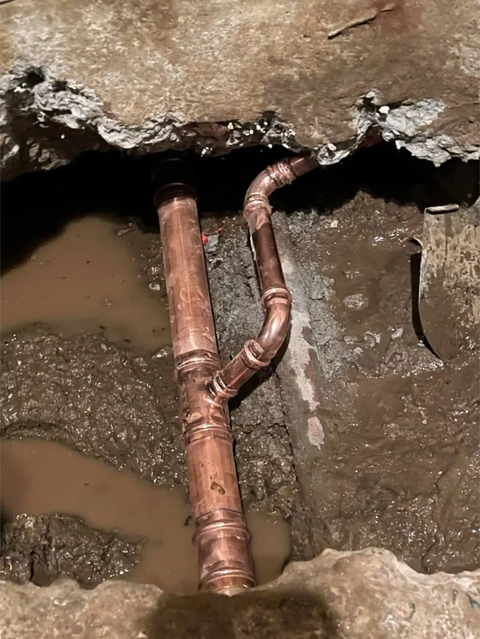Exposed copper plumbing pipes in a muddy trench beneath a broken floor.