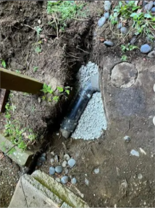 Exposed drainage pipe in a dirt trench beside a concrete walkway and scattered stones