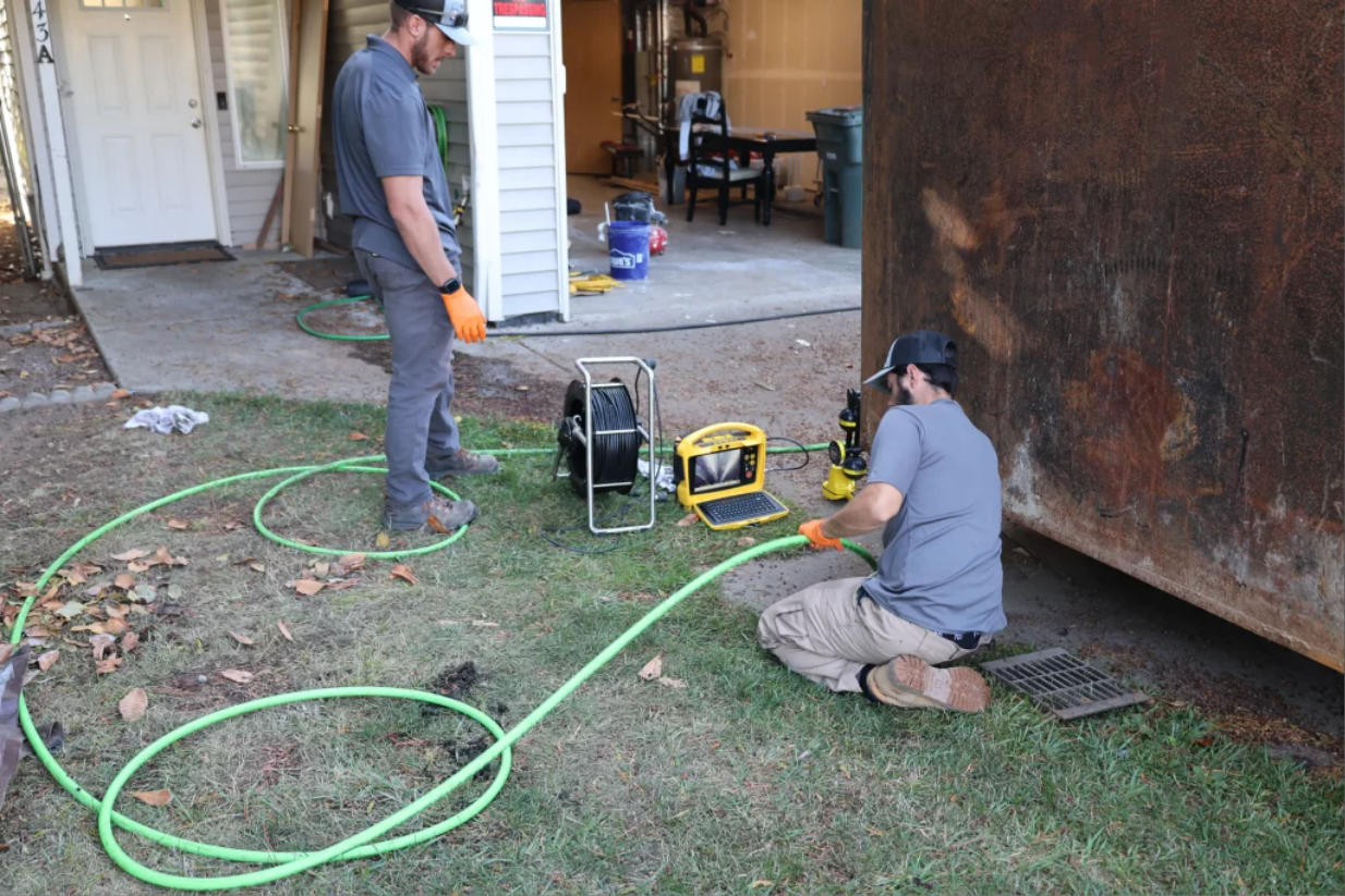 Two plumbing technicians with a camera reel, monitor and hose beside a garage