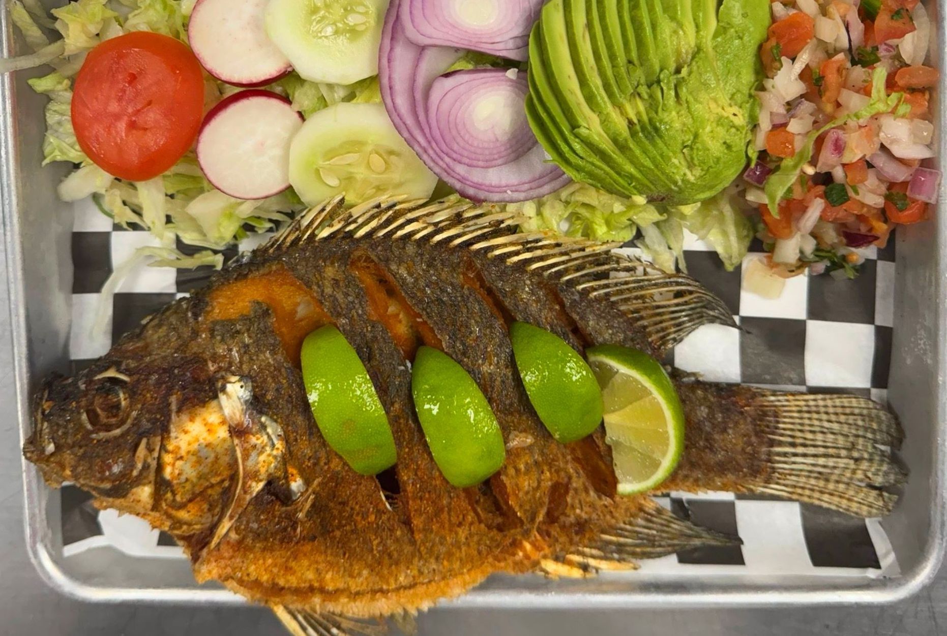 Fried whole fish with lime, avocado, and vegetables in a tray.