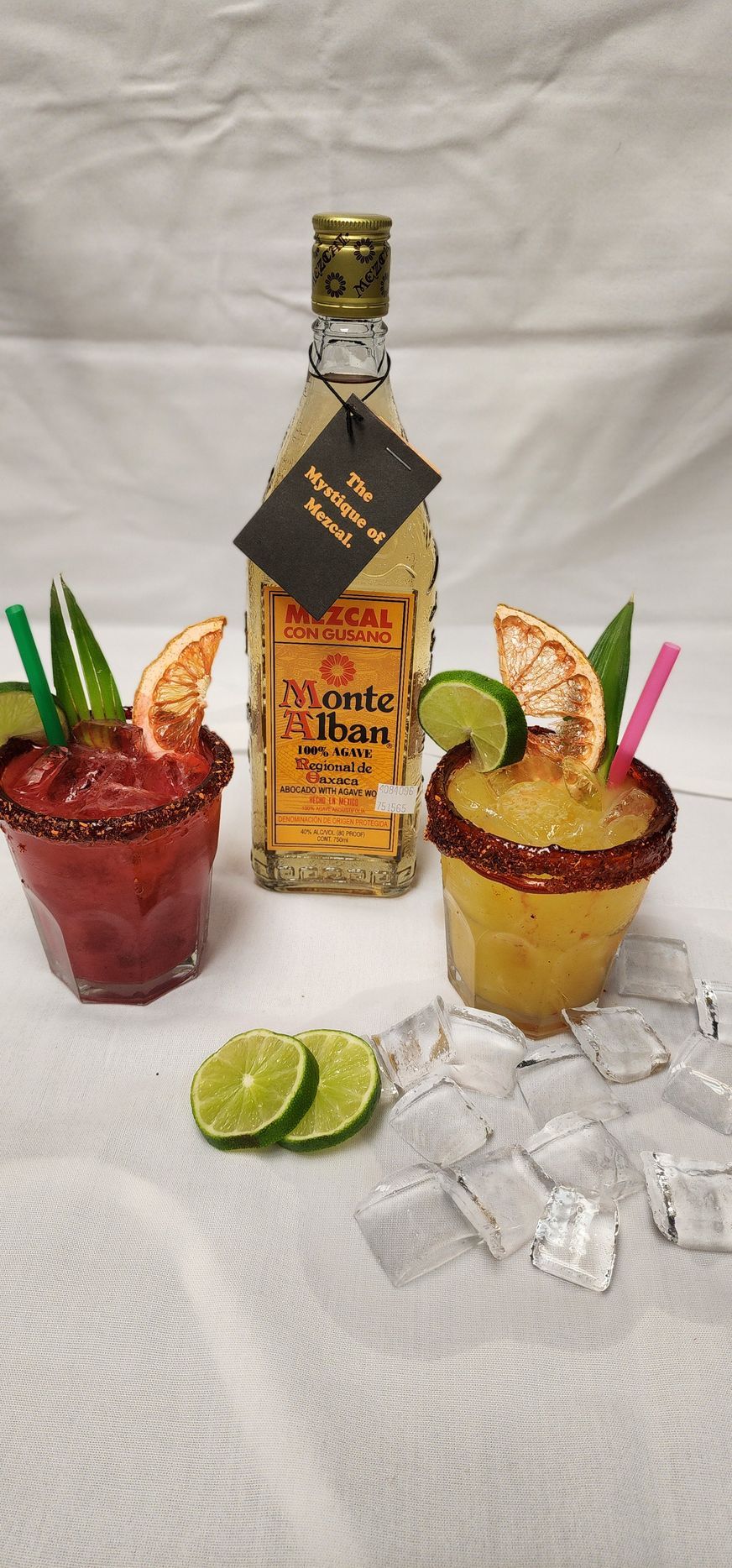 Two cocktails with a bottle of tequila on a white surface, garnished with lime and orange slices.