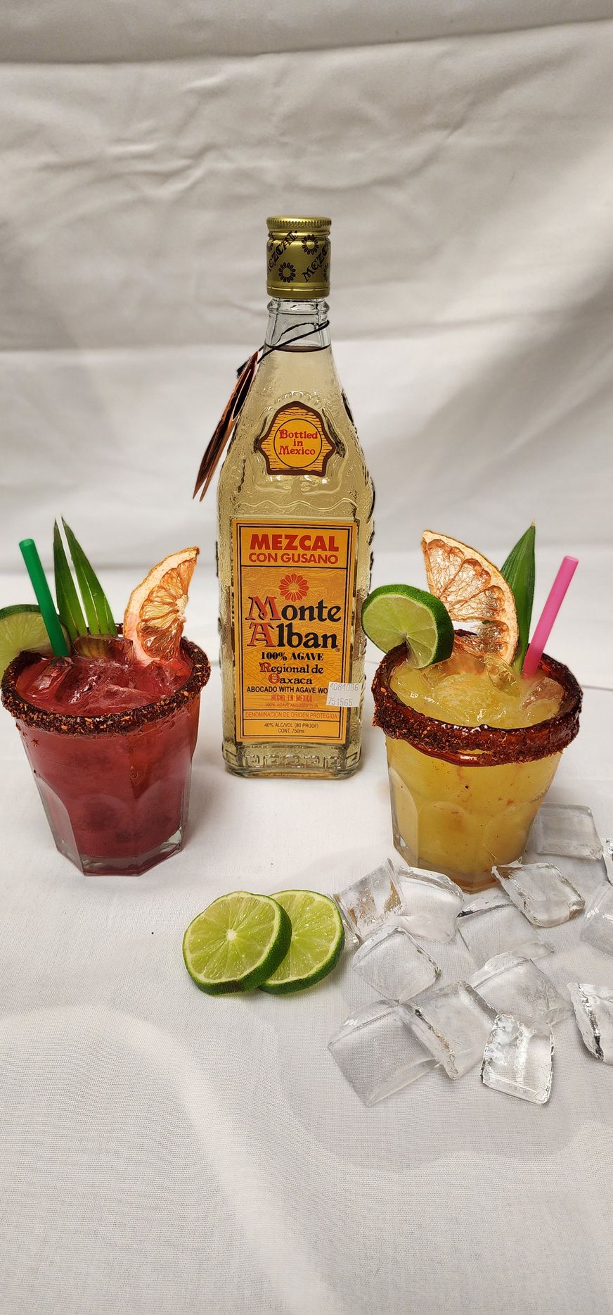 Bottle of Monte Alban mezcal with two cocktails, lime slices, and ice cubes on a white background.