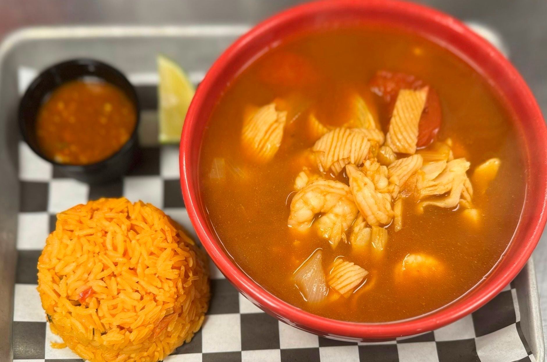 Seafood soup with rice and sauce on a checkered tray. Red bowl, orange rice, and a lime wedge.