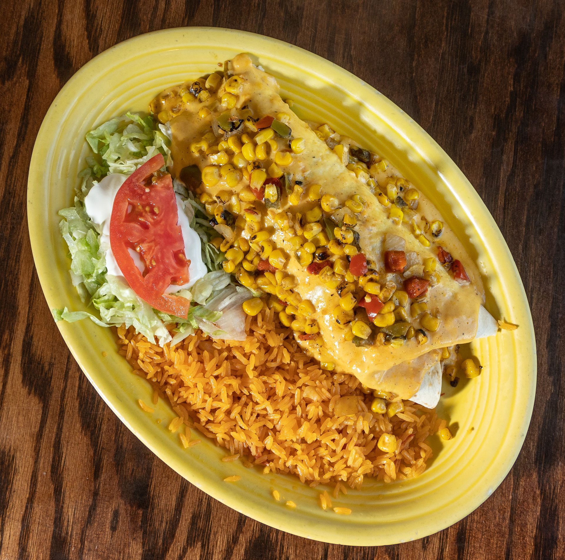 Enchiladas with corn topping, rice, lettuce, tomato, and sour cream on a yellow plate.