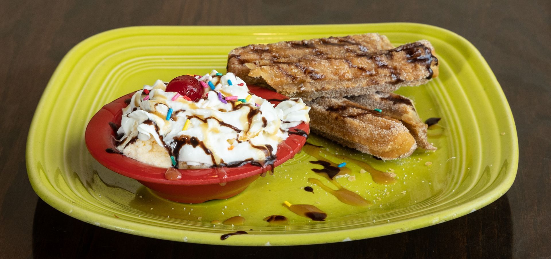 A dessert on a yellow plate. It has ice cream in a red bowl, churros, and chocolate syrup.