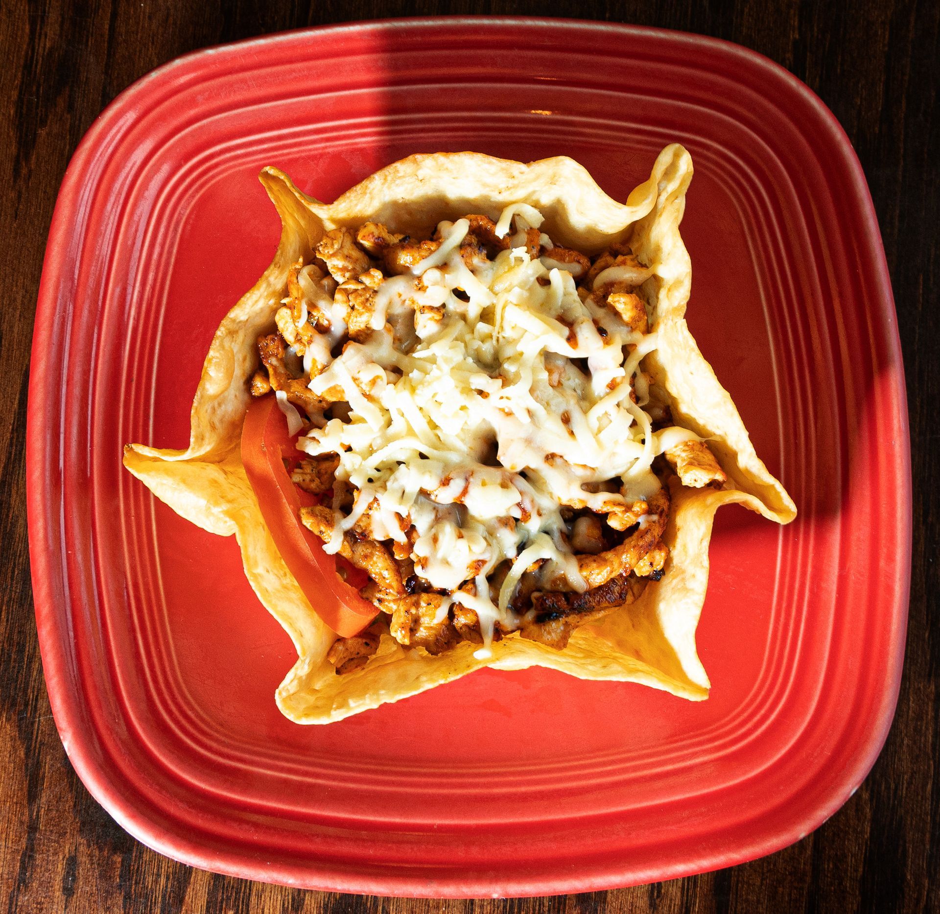 Taco salad in a crispy shell, topped with shredded cheese and a slice of tomato on a red plate.