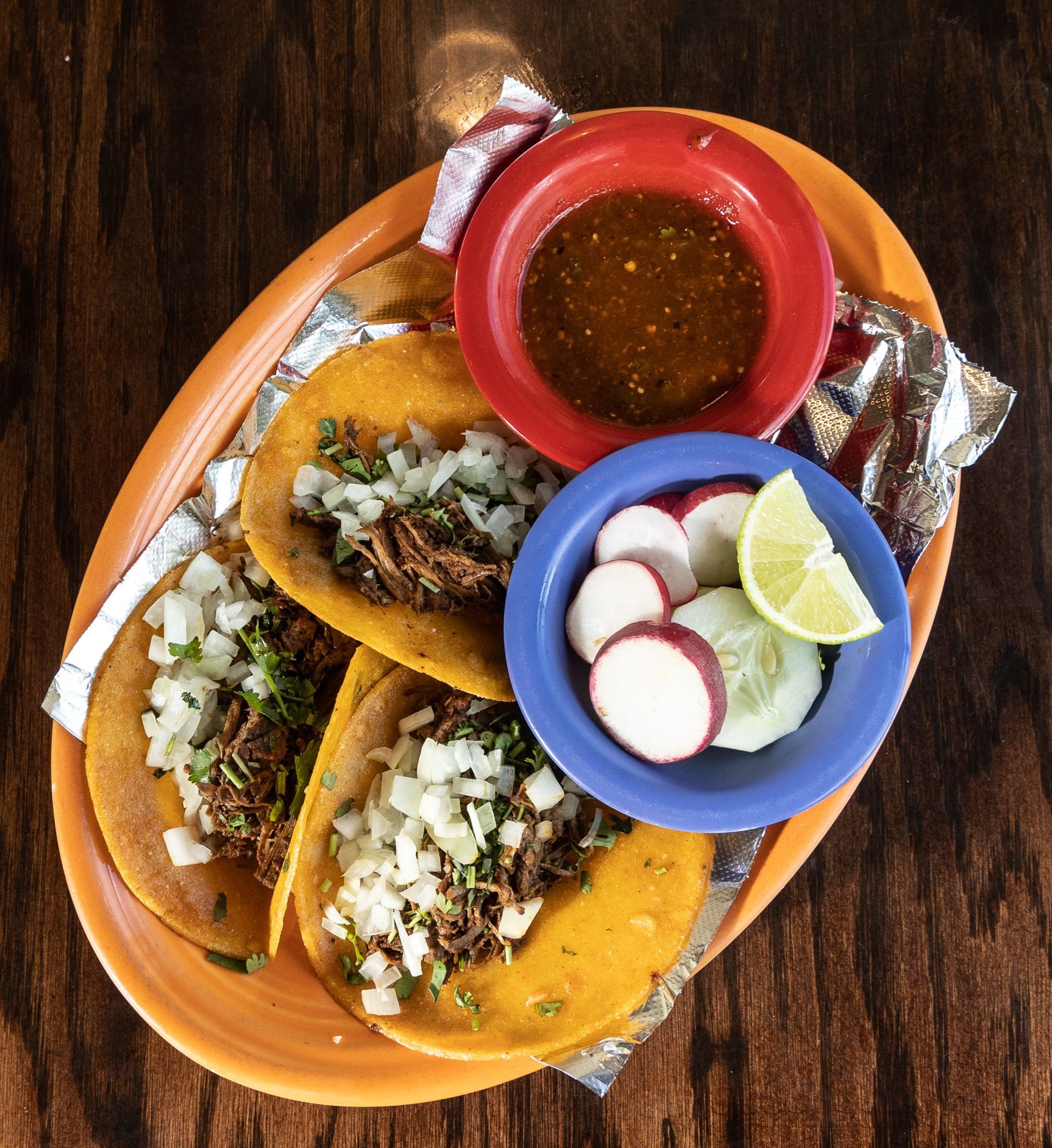 Three tacos with onion and meat, salsa, radishes, and lime on a plate.