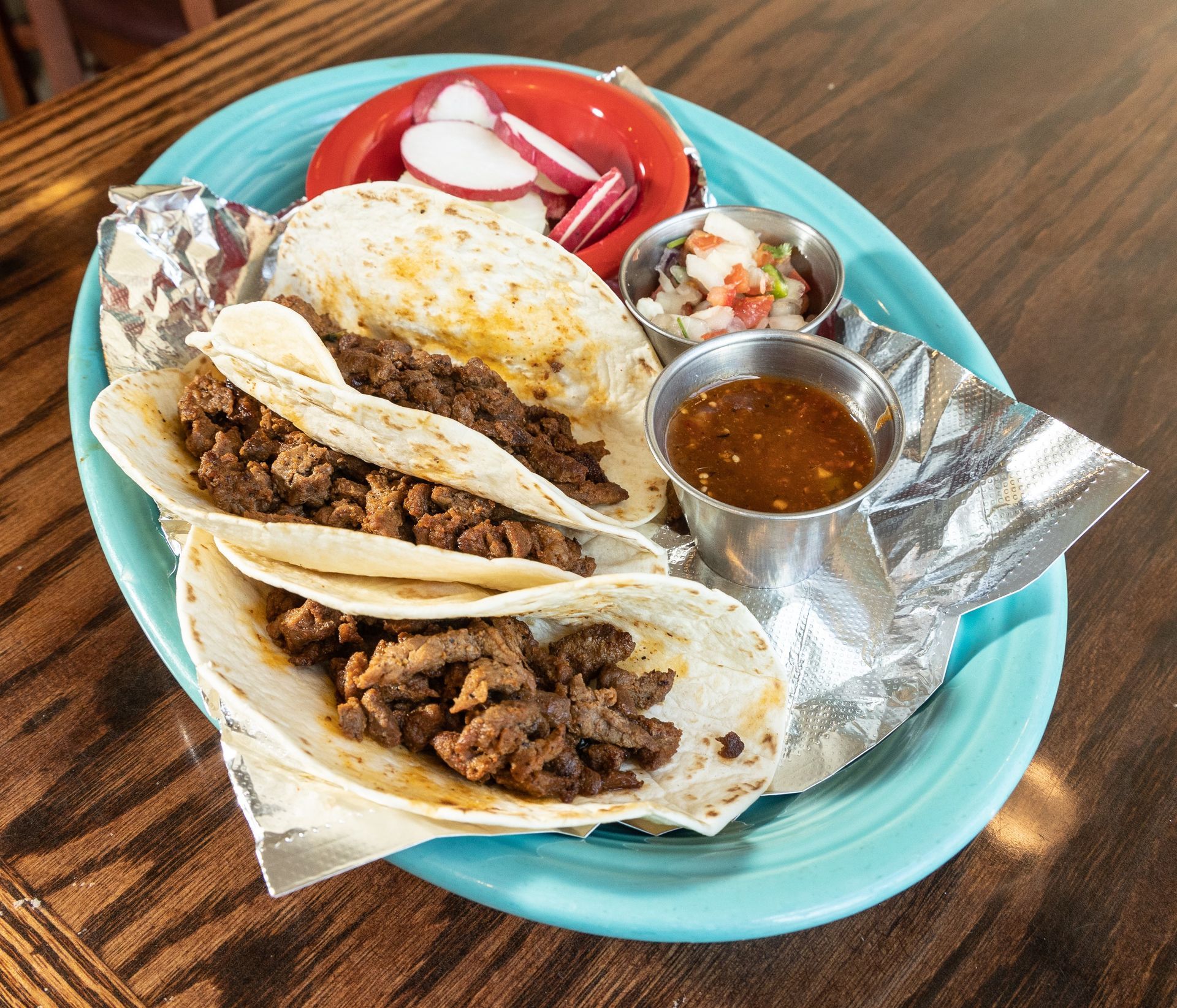 Tacos with meat, served with salsa, radishes, and sauce on a teal plate.