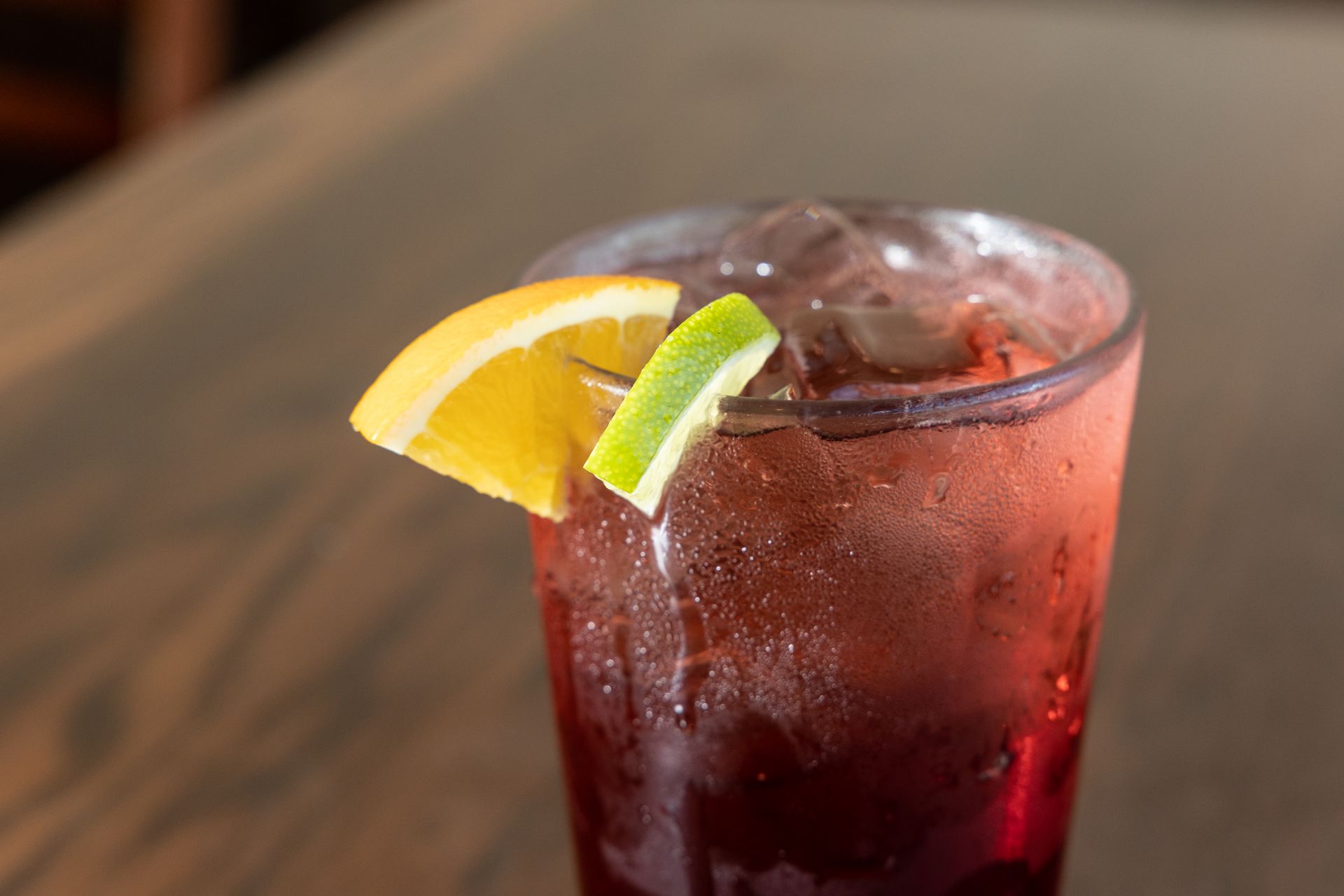 A glass of reddish-brown drink with ice, garnished with orange and lime slices, on a wooden surface.