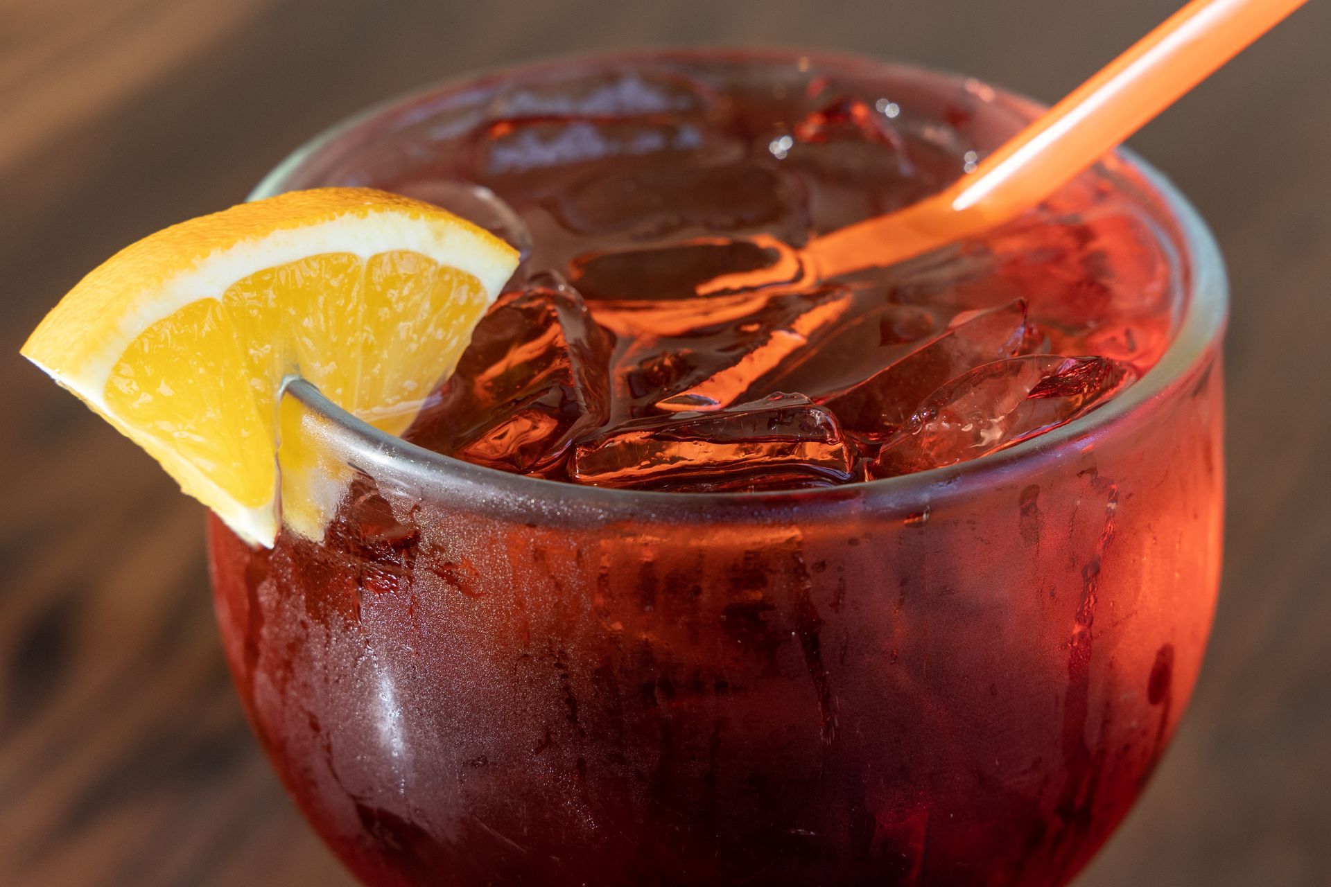 Glass of reddish-brown drink with ice, orange slice garnish, and orange straw.