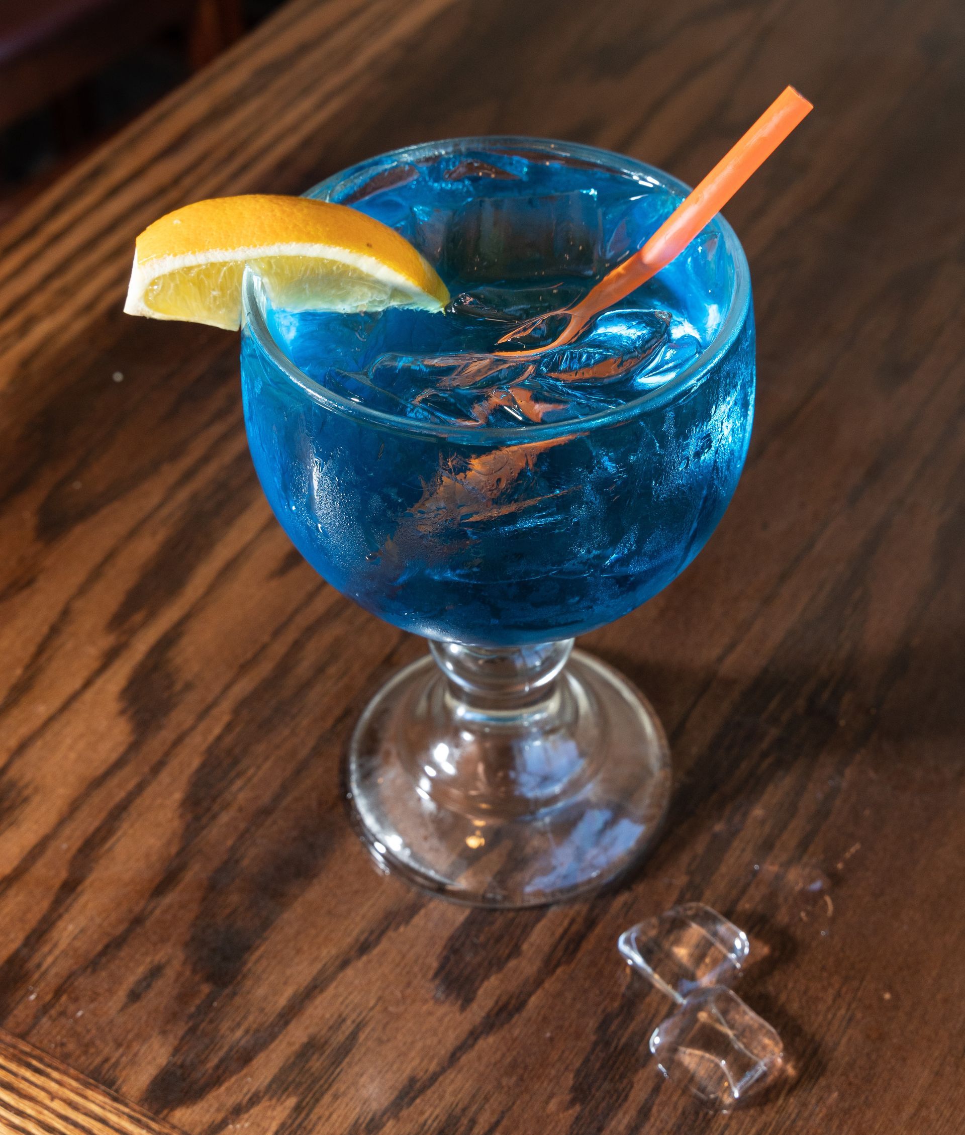 Blue cocktail in a goblet, lemon garnish, straw, on a wooden table.