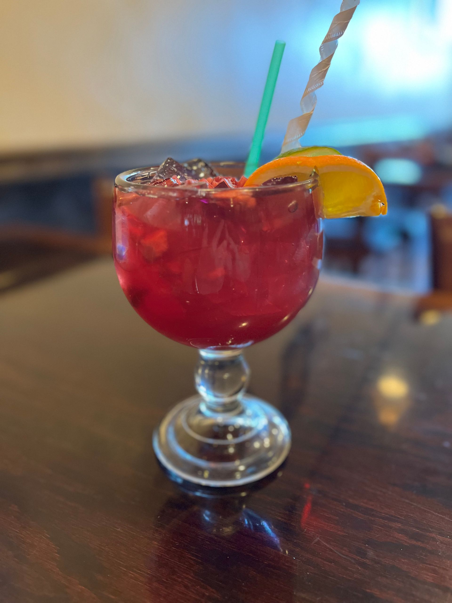 A red cocktail in a stemmed glass with ice, orange slice, and two straws, on a wooden table.