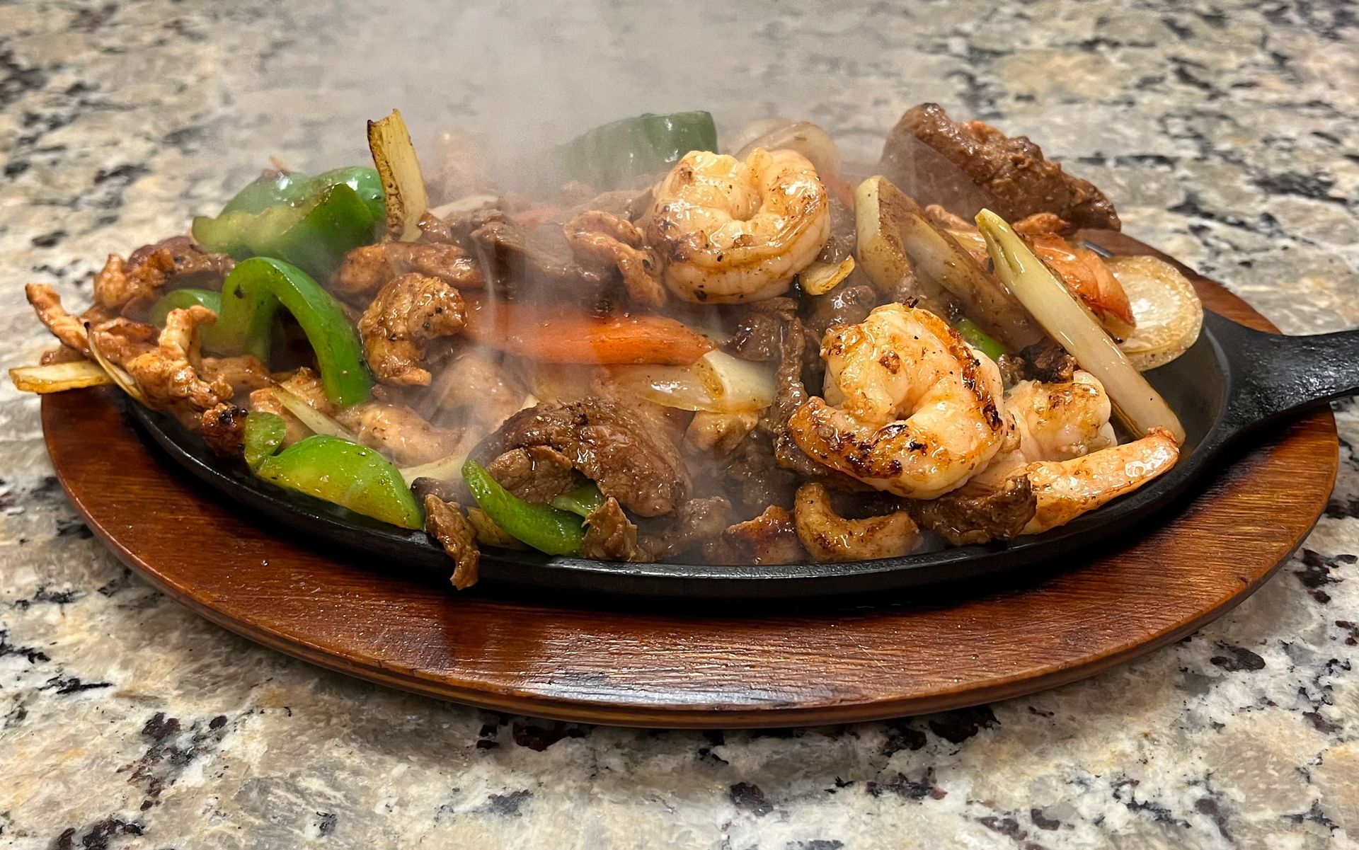 Sizzling skillet of fajitas: shrimp, chicken, vegetables. Steam rises. Served on a wooden plate.