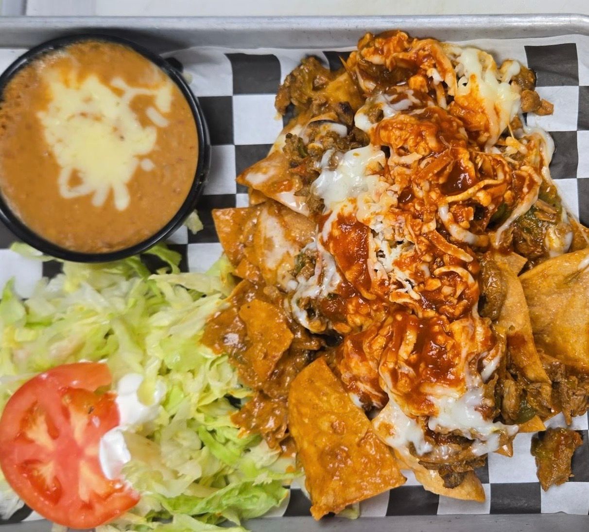 Nachos with beans, lettuce, and a tomato slice on a checkered tray.