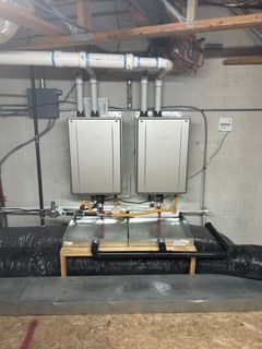 Two wall-mounted boilers with white pipes and a black insulated tank in a basement utility room