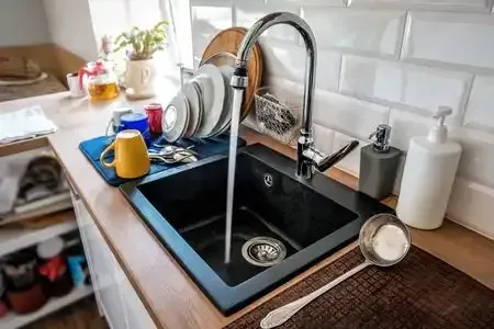 Kitchen sink with running faucet, black basin, blue mat, yellow mug, and white tiled backsplash Kitchen sink with running faucet, black basin, blue mat, yellow mug, and white tiled backsplash