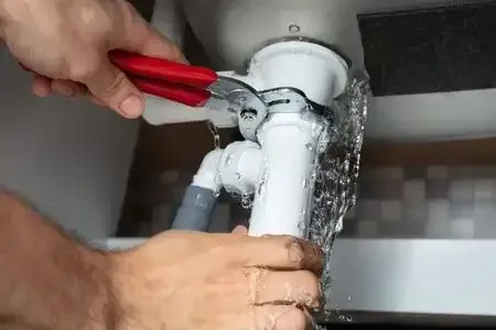 Hands using red pliers to tighten a leaking white pipe under a sink.
