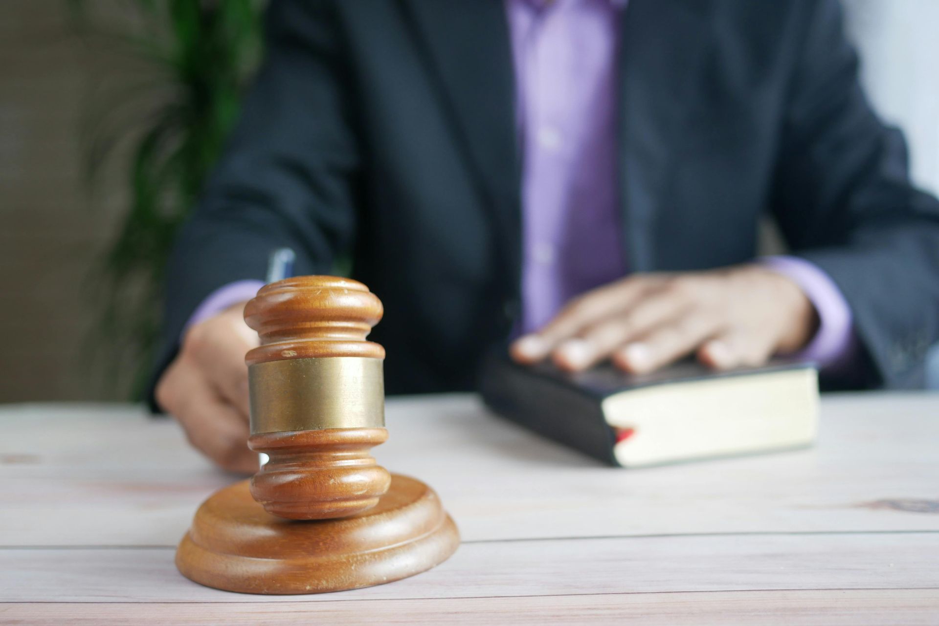 Wooden gavel on desk with person's hand on book, wearing a suit.