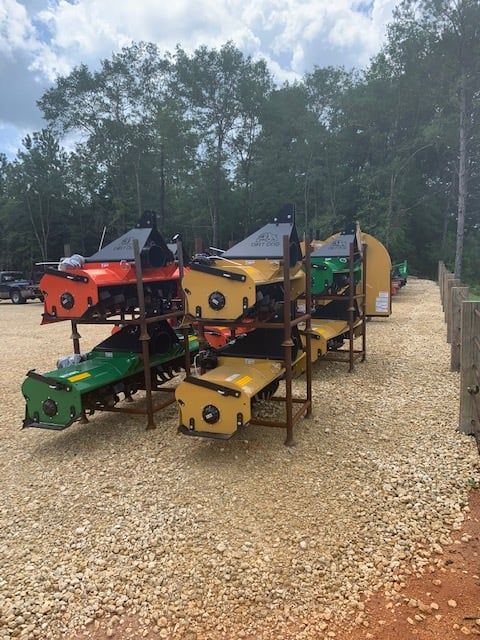 Several colored tractor attachments on metal shelving on a gravel lot, trees in background.