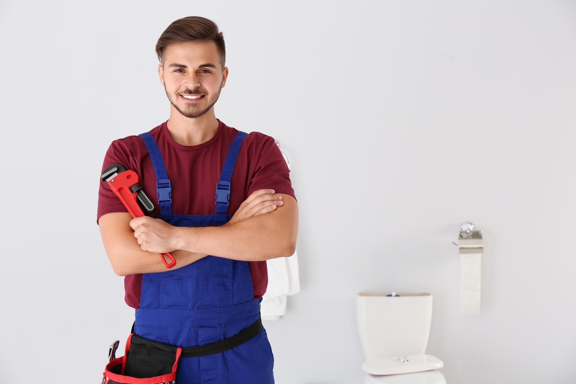 Smiling plumber in blue overalls holding a wrench, standing in a bathroom.