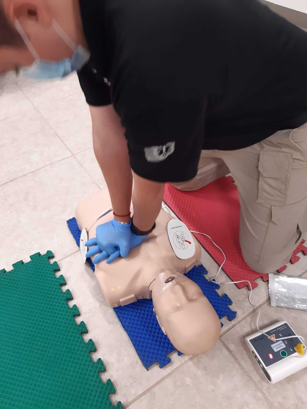 Persona con camisa negra, guantes azules, arrodillada, realizando RCP a un maniquí.