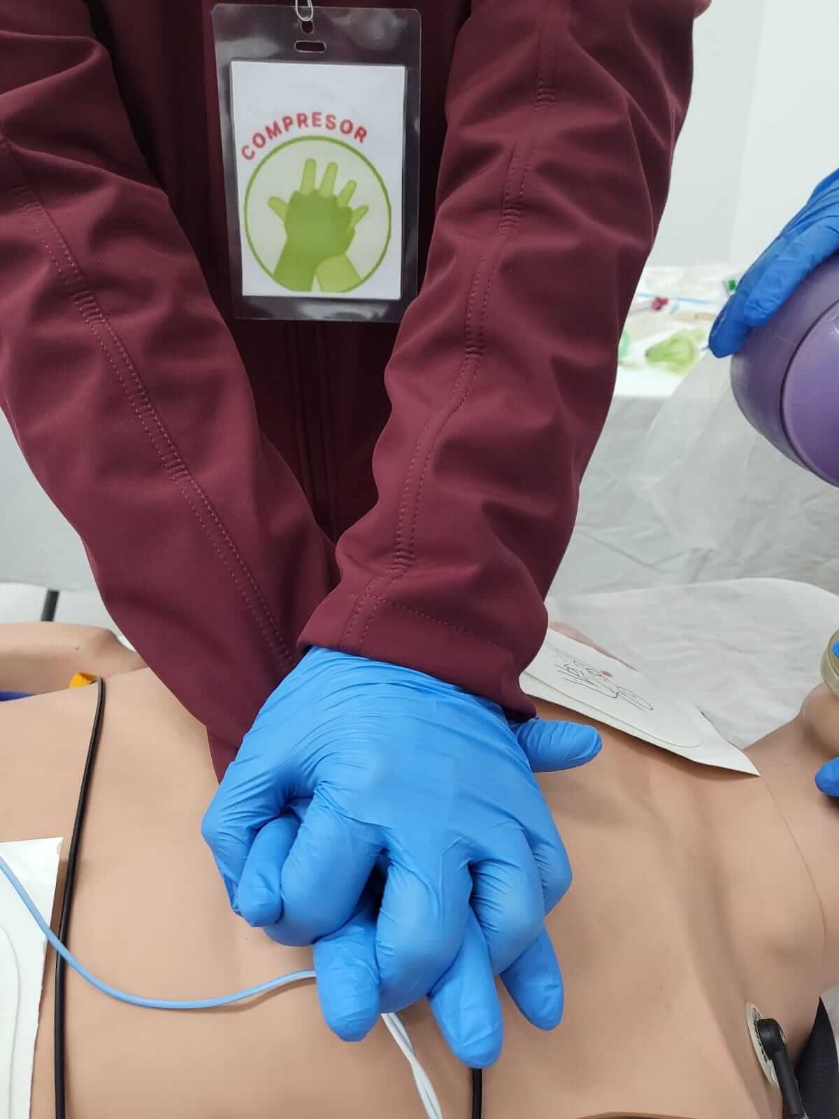 Persona realizando RCP en un maniquí, con las manos entrelazadas y guantes azules. La placa indica