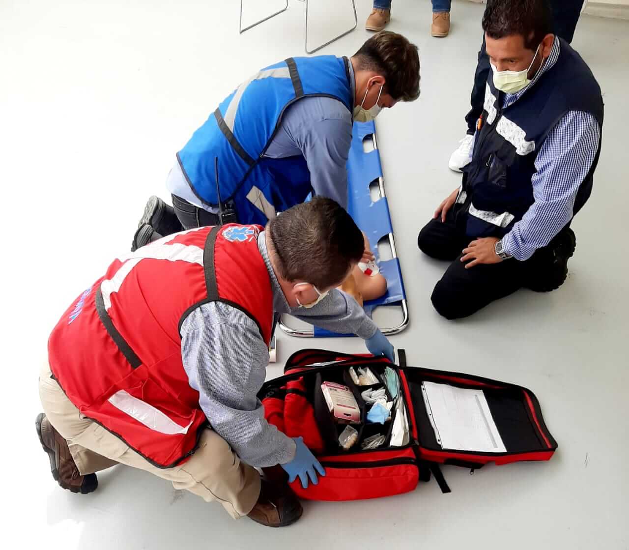 Tres personas con chalecos practican primeros auxilios en un maniquí, con una bolsa médica abierta y una camilla.