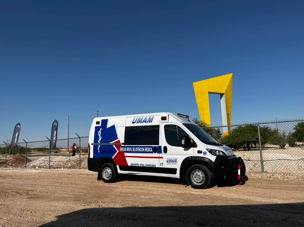 Ambulancia blanca con detalles azules y rojos estacionada al aire libre, con una escultura amarilla al fondo.