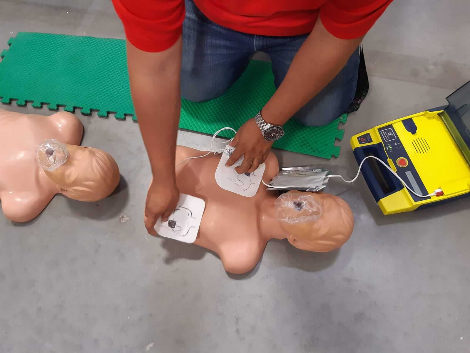 Persona colocando parches de DEA en un maniquí de entrenamiento tendido en el suelo. Cerca hay un DEA amarillo.