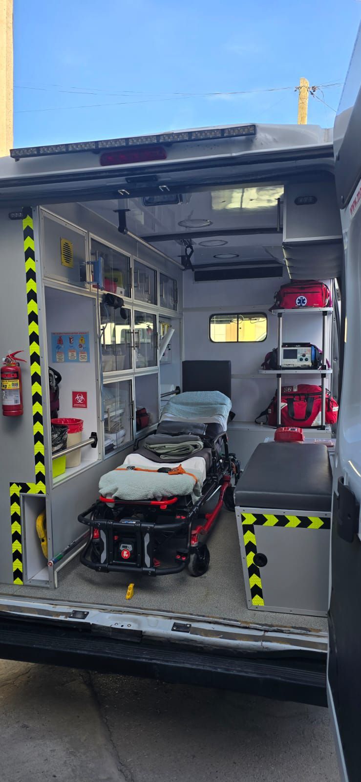 Interior de una ambulancia con camilla, equipo médico y compartimentos de almacenamiento. Cinta de peligro negra y amarilla.