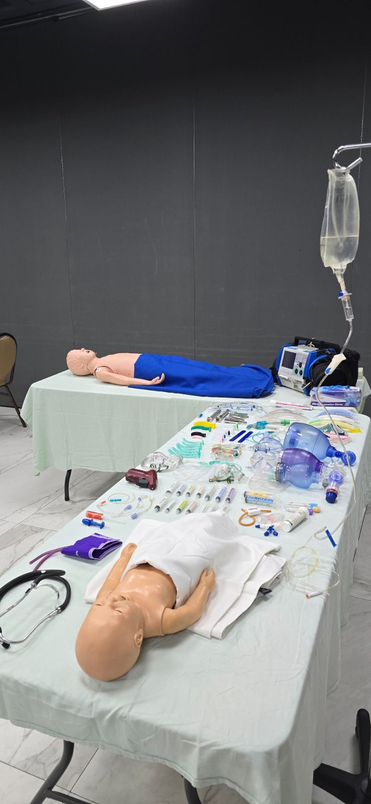 Una sala de formación médica con maniquíes médicos adultos y bebés sobre mesas rodeadas de suministros.