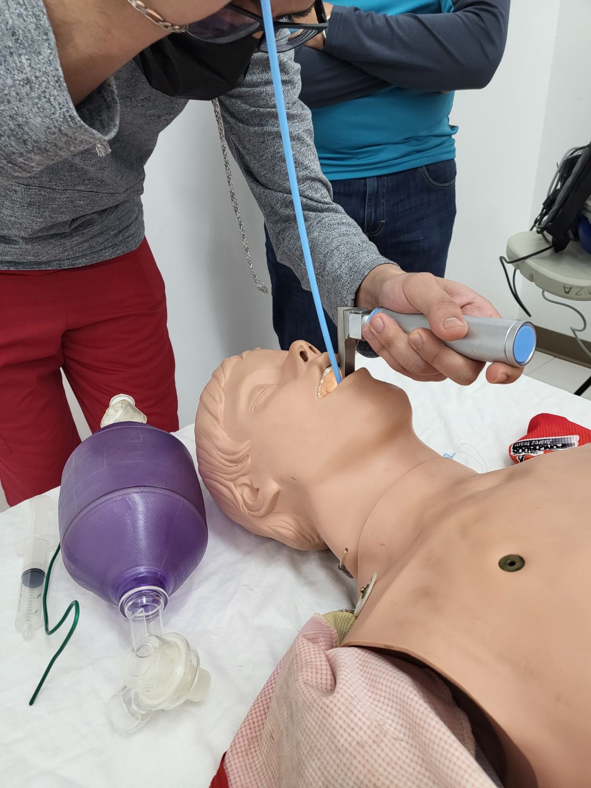 Maniquí intubado durante una formación médica, con personas usando equipo. Bolsa morada y tubo azul.