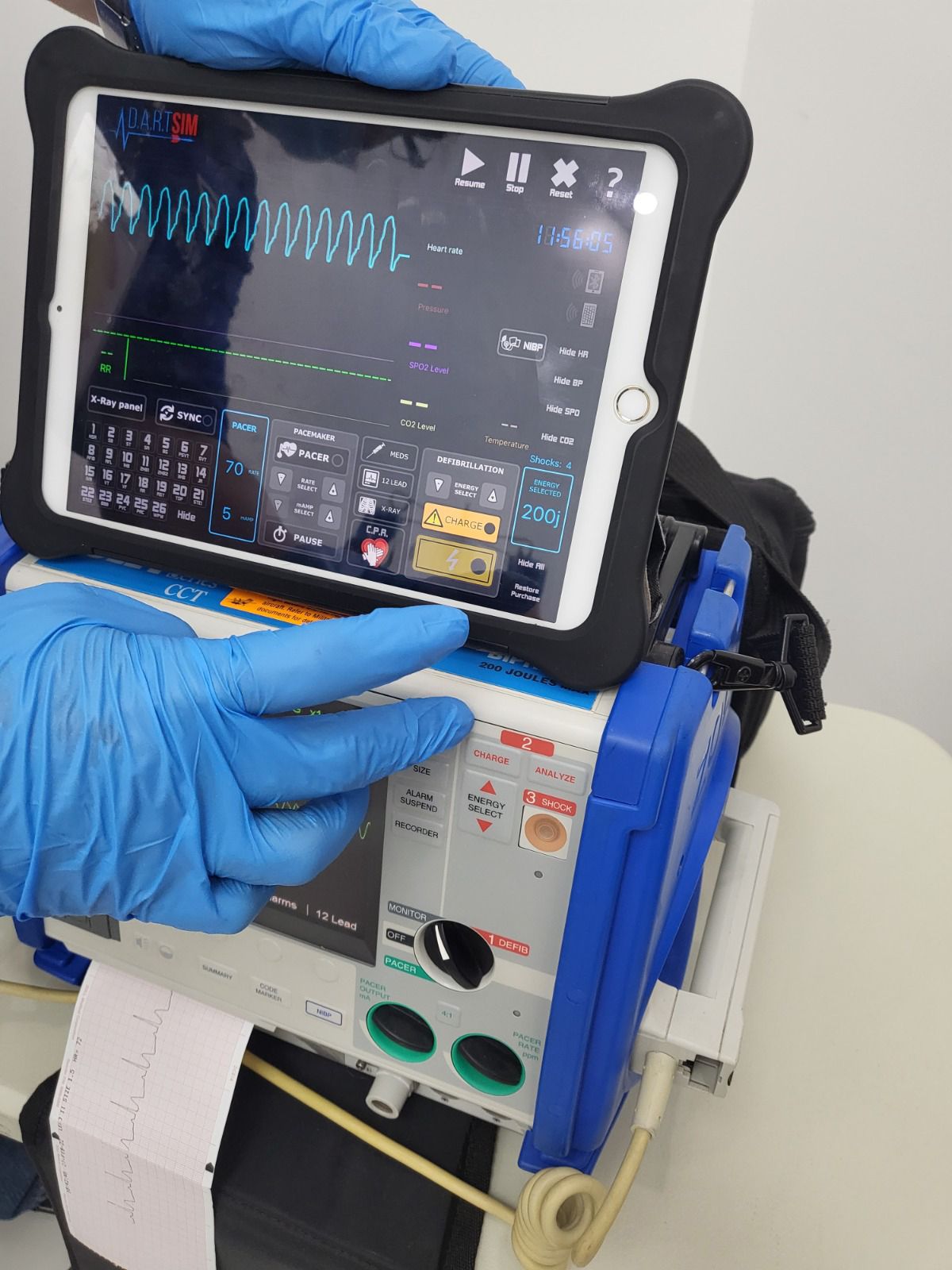 Persona que usa guantes azules y sostiene una tableta que muestra lecturas del ritmo cardíaco en un dispositivo médico de emergencia.
