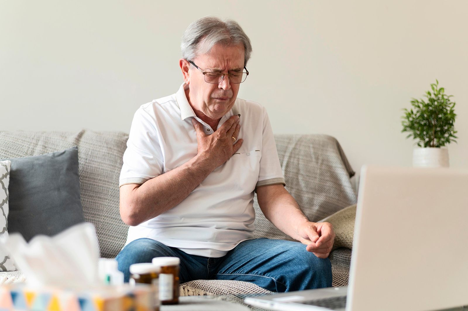 Hombre agarrándose el pecho mientras está sentado en un sofá, mirando una computadora portátil con expresión enfermiza.