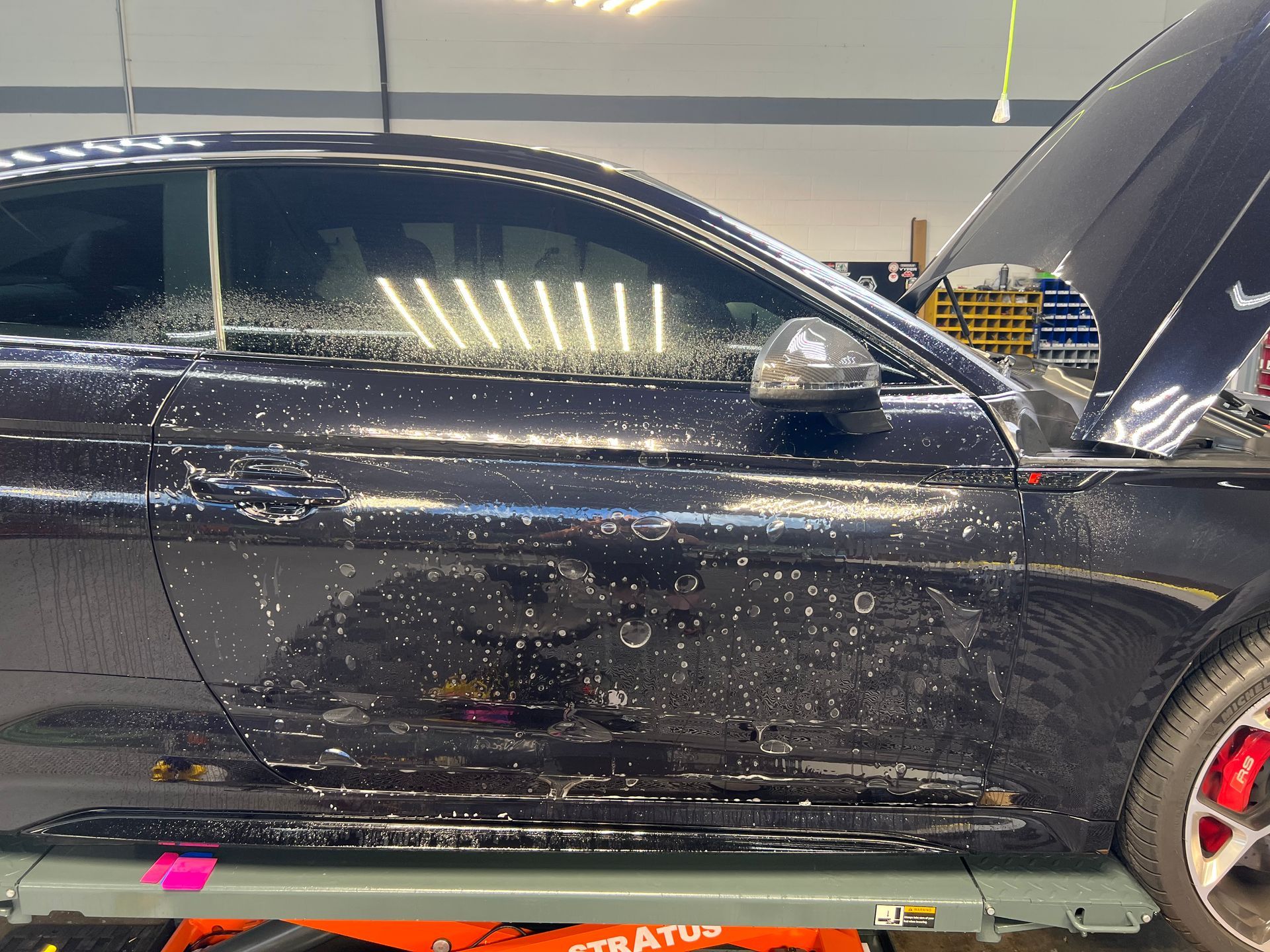 Black car with open hood being worked on, inside a shop. The car has wet spots on its side.
