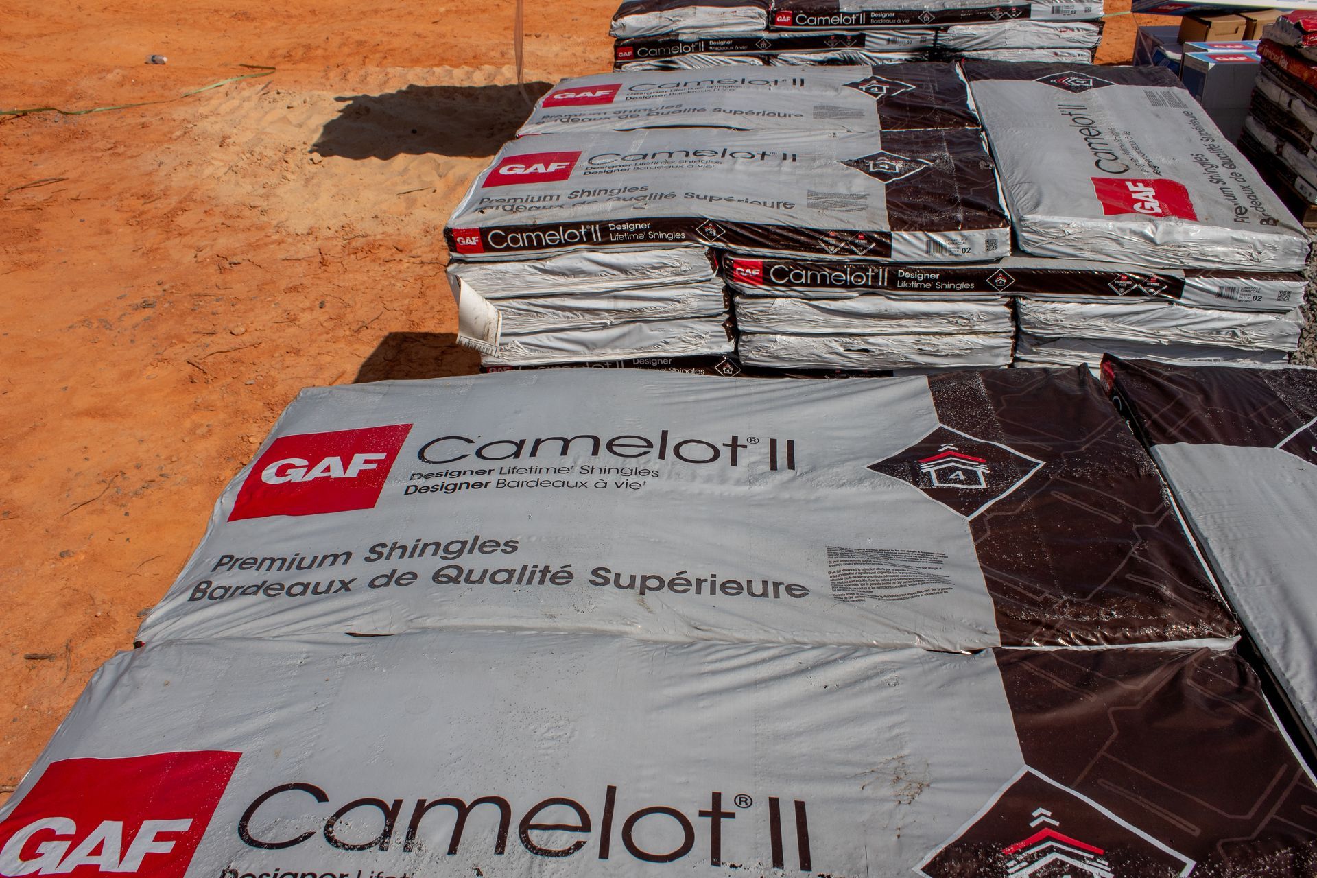Stacks of GAF Camelot II roofing shingles on a dirt surface, ready for installation.
