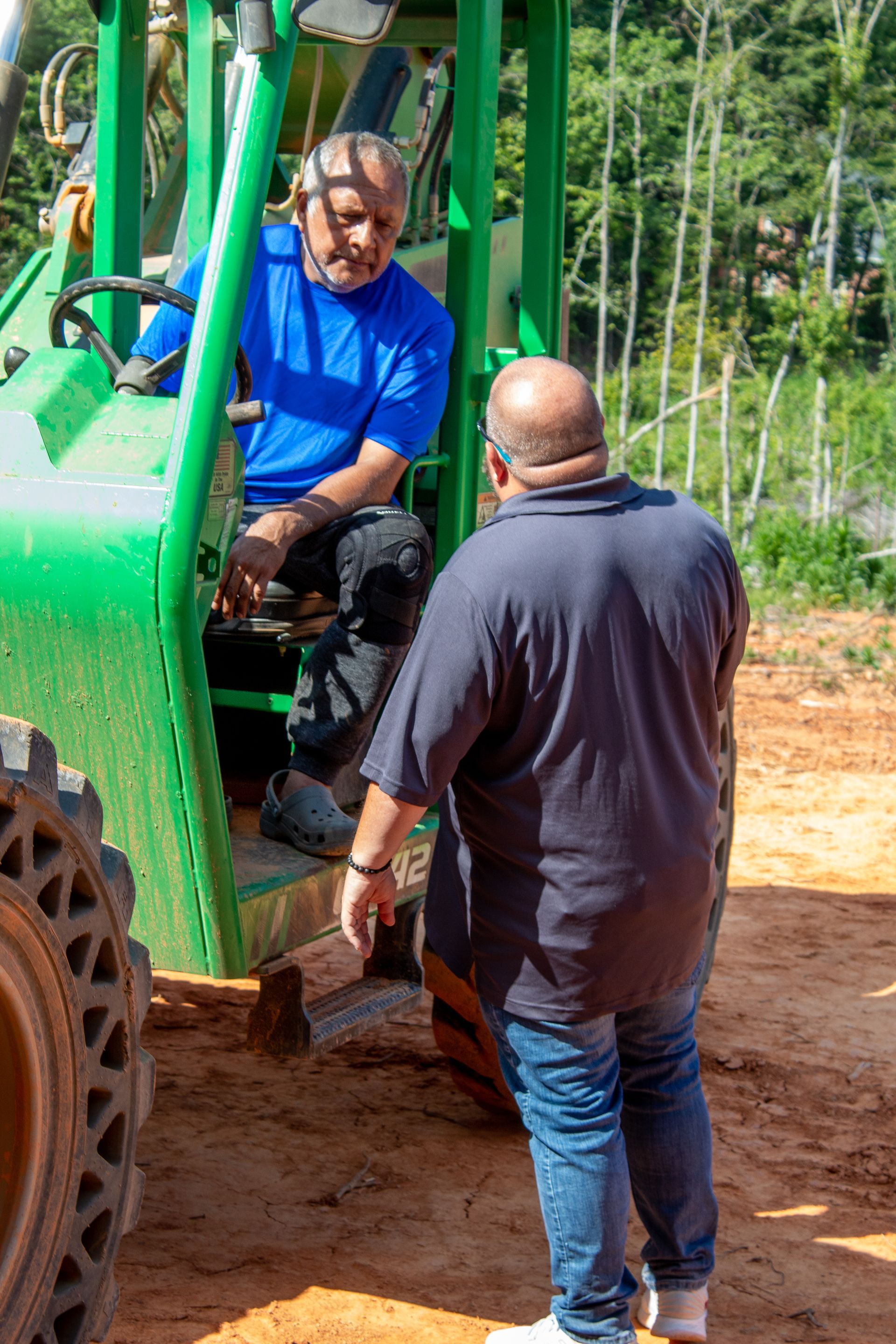 Two men talking by a green construction vehicle. One man sits inside, the other stands outside.