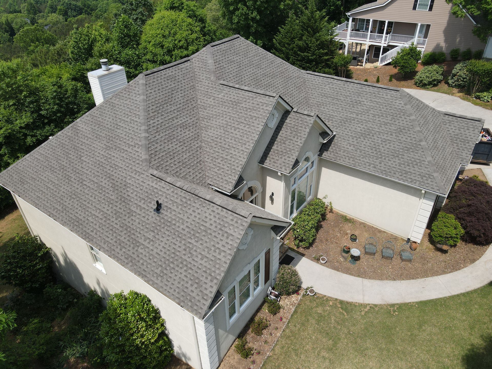 Aerial view of a beige house with a gray shingle roof, surrounded by green trees and grass on a sunny day.