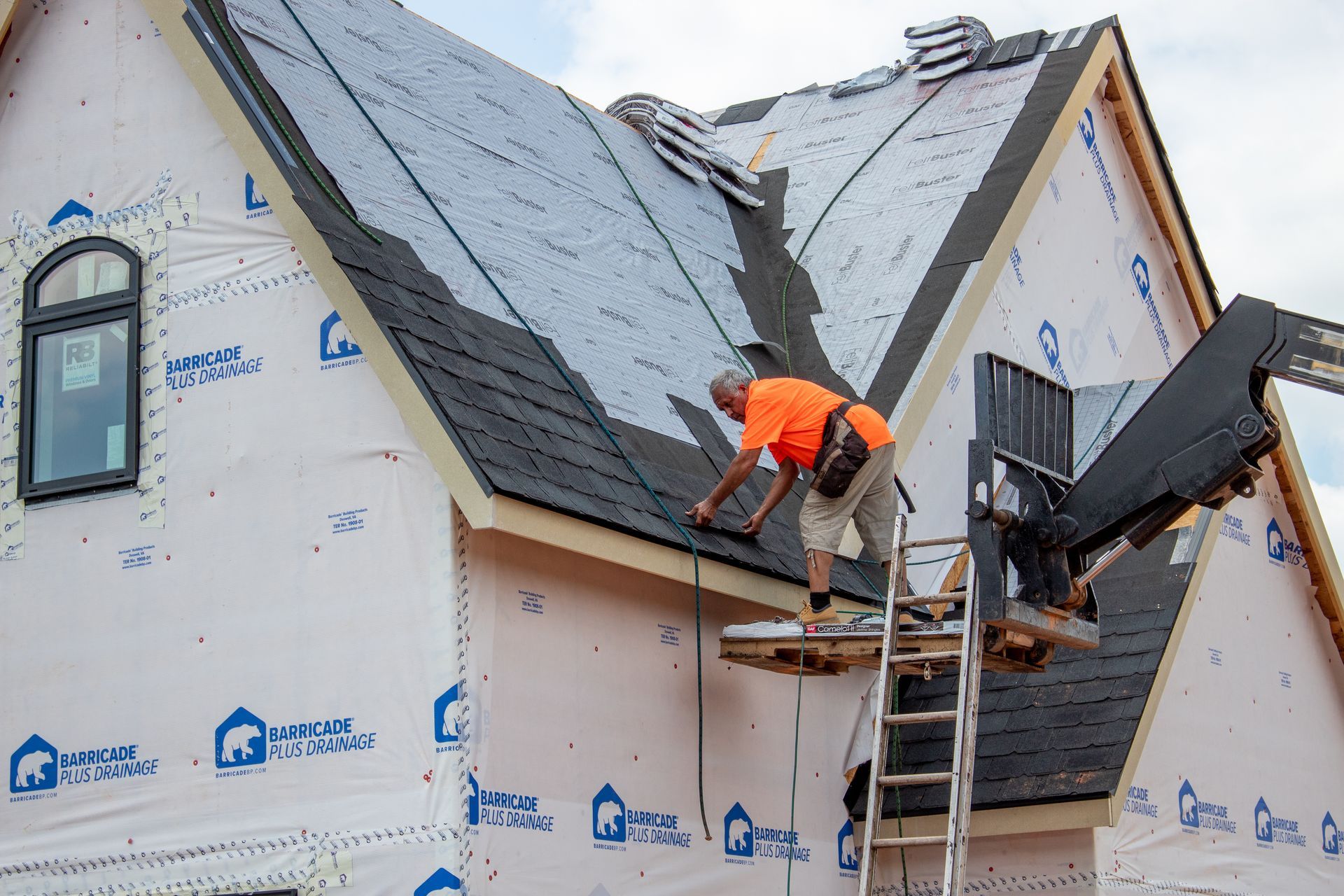 Roofer on a ladder installing shingles on a house roof, using a boom lift.