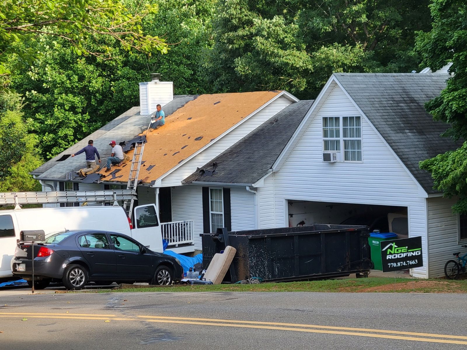 House with roof partially torn off; workers on roof; dark car, dumpster, and trees in the background.