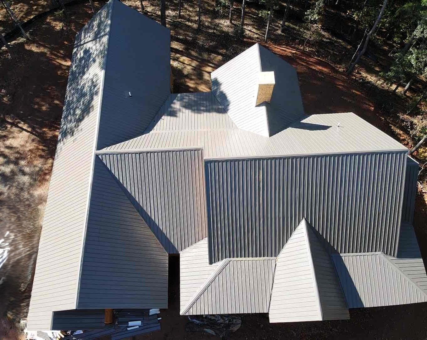 Overhead view of a modern house with a complex, light gray corrugated metal roof and chimney, surrounded by trees.