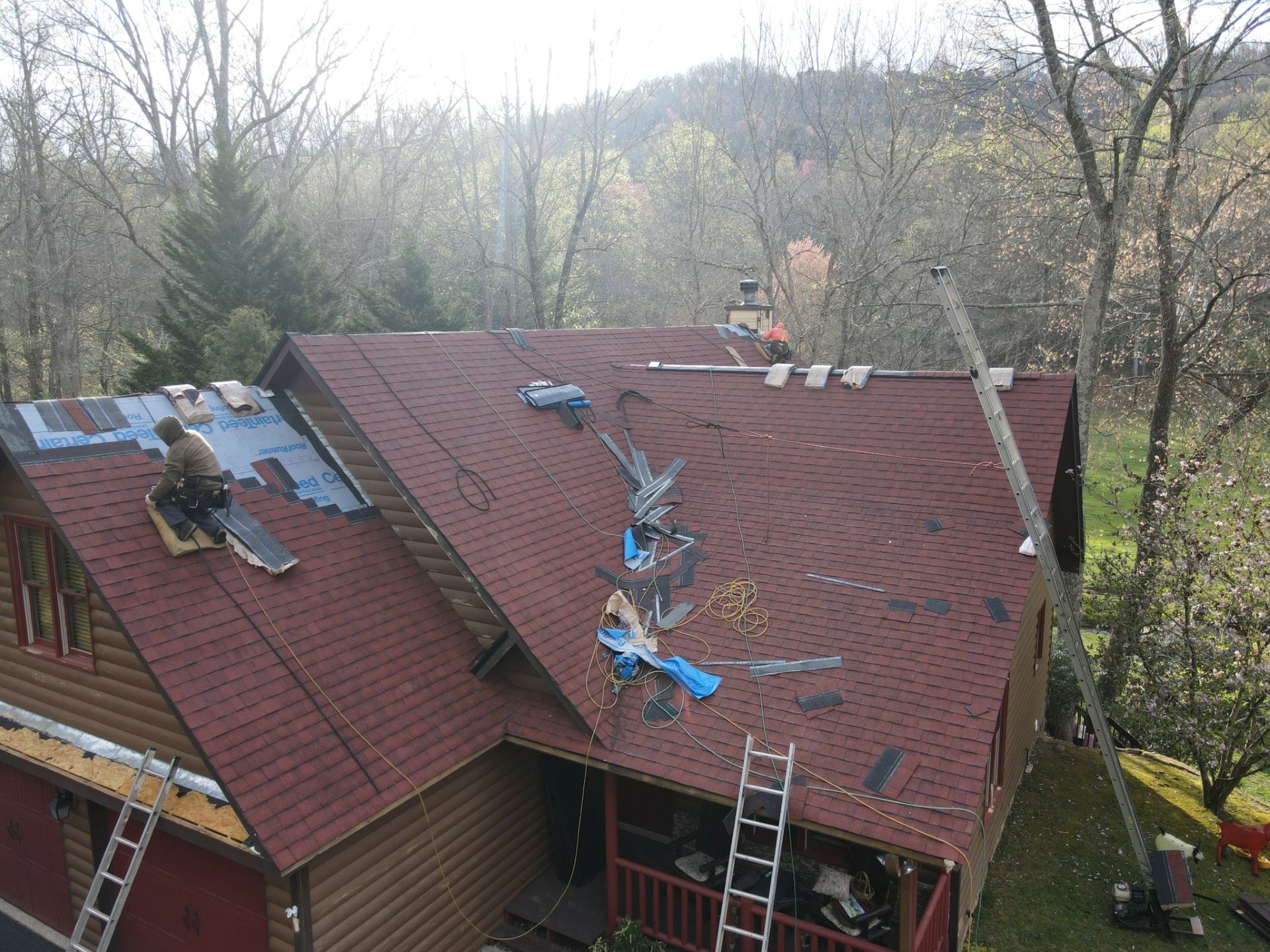 Workers installing shingles on a house roof; fall foliage in the background. Ladders and tools are present.