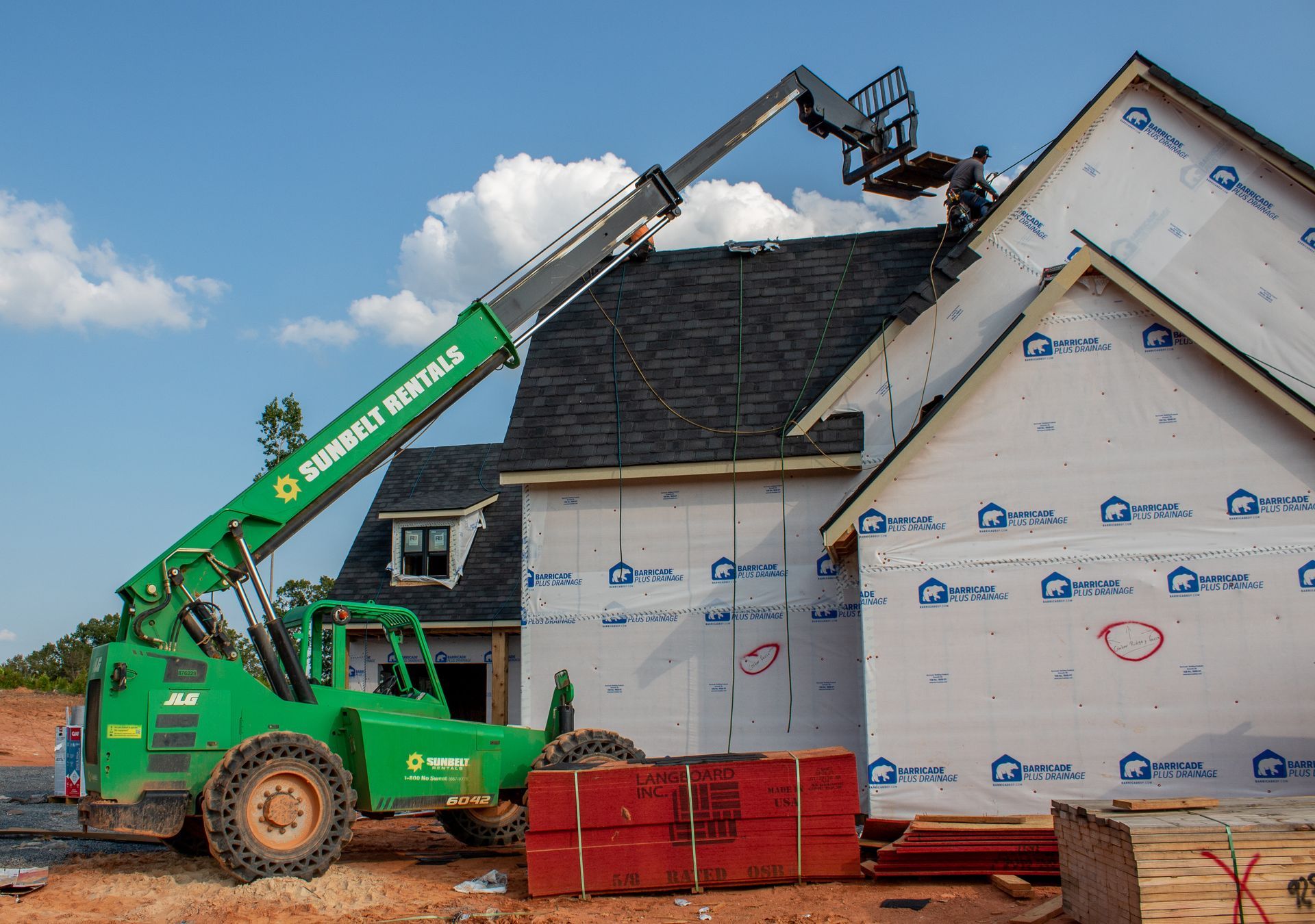 Construction worker on roof using a telehandler to install siding. Green lift, wood and blue-wrapped building.