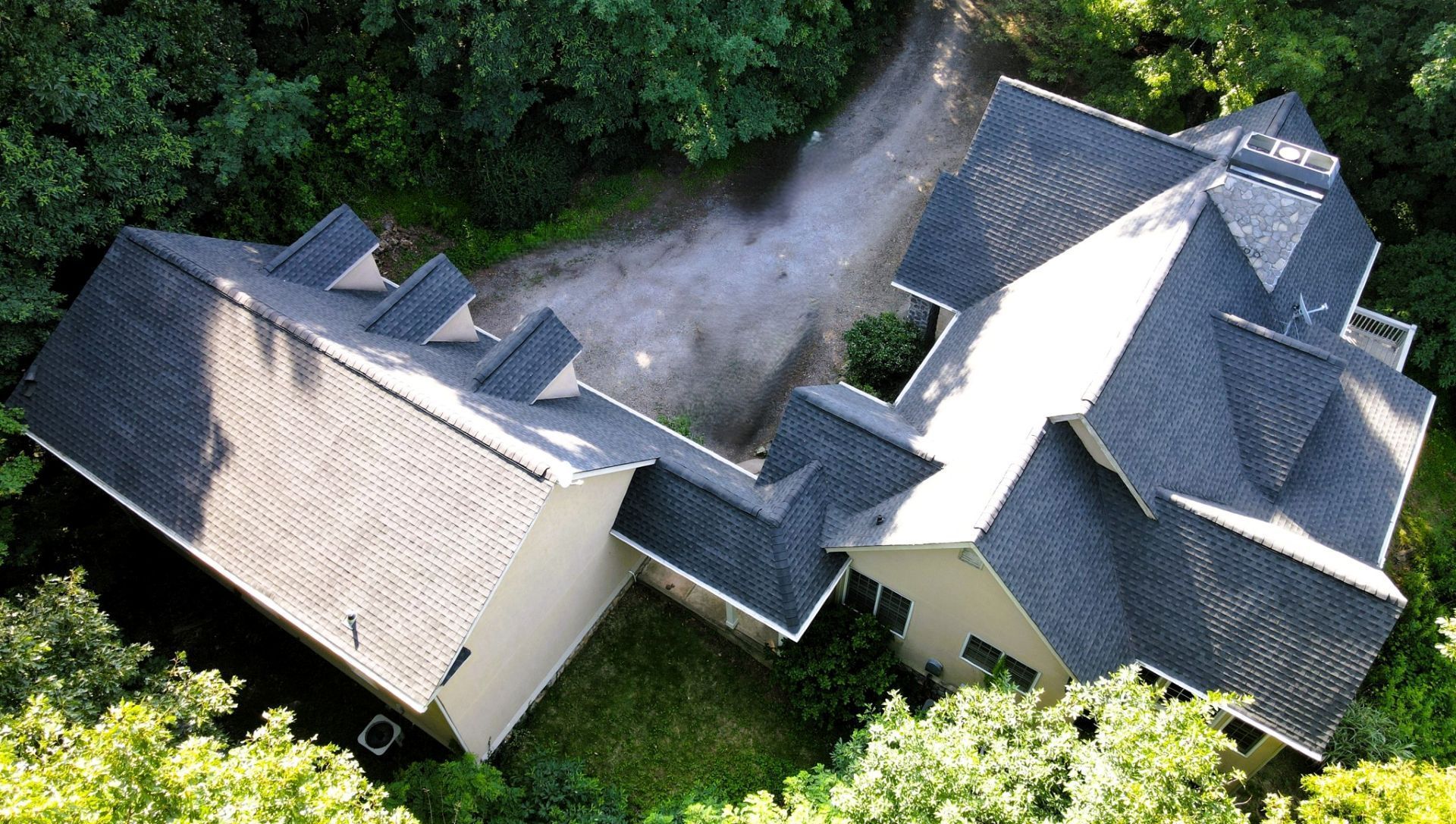 Overhead view of a house with a complex, dark-roofed structure, surrounded by green trees and a gravel driveway.