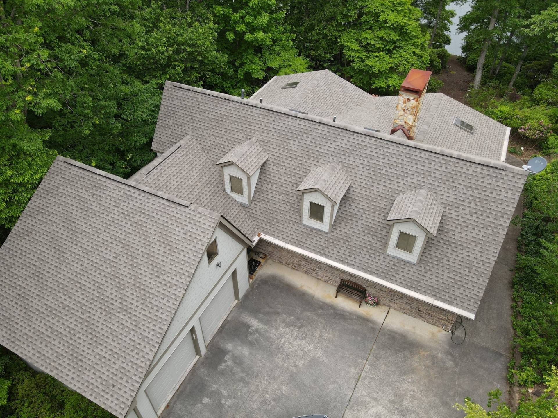 Aerial view of a house with gray shingle roof, dormers, and a chimney, surrounded by green trees.