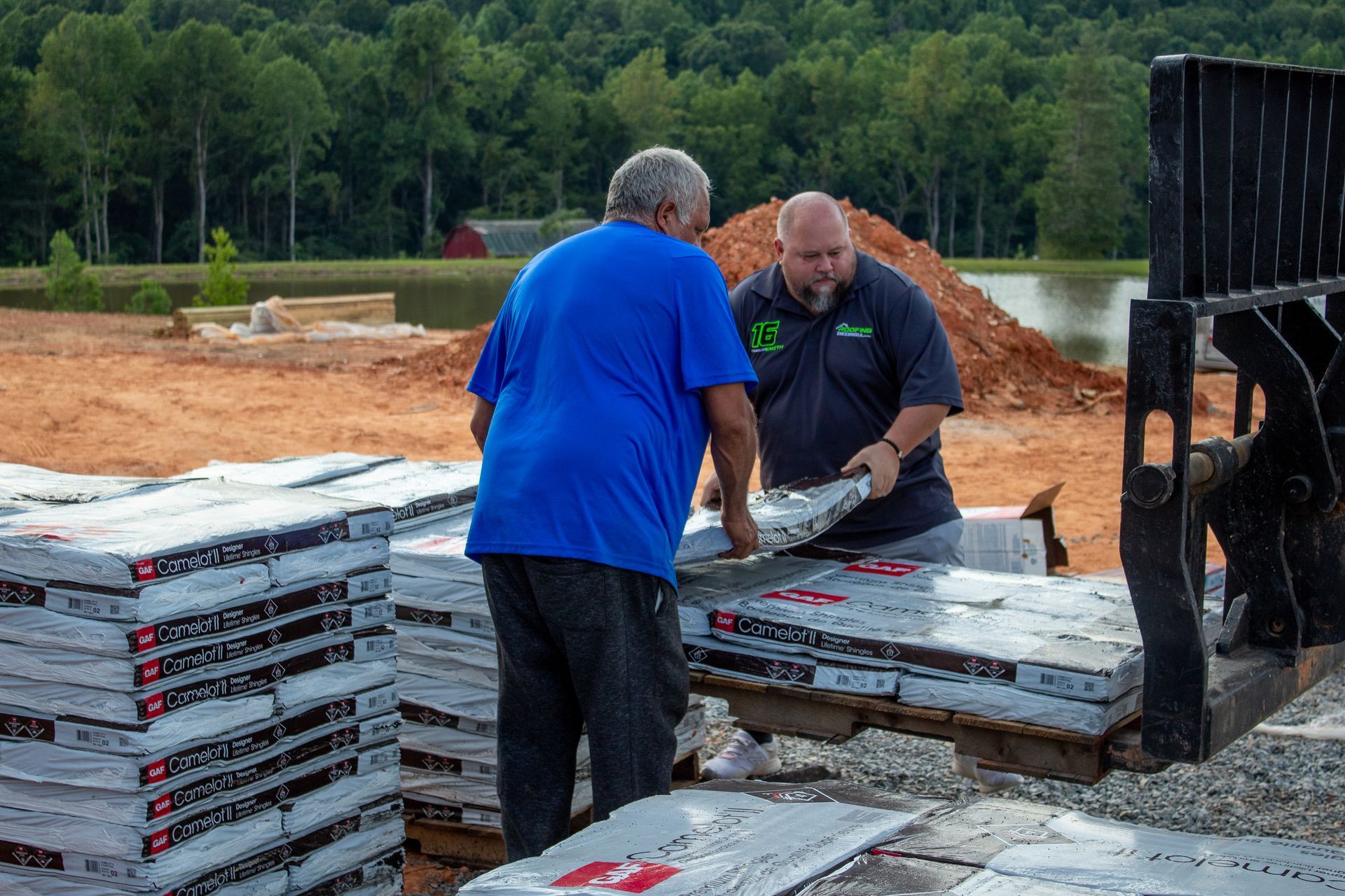 Two people loading materials from a pallet. Outdoors, near a pond and construction site.