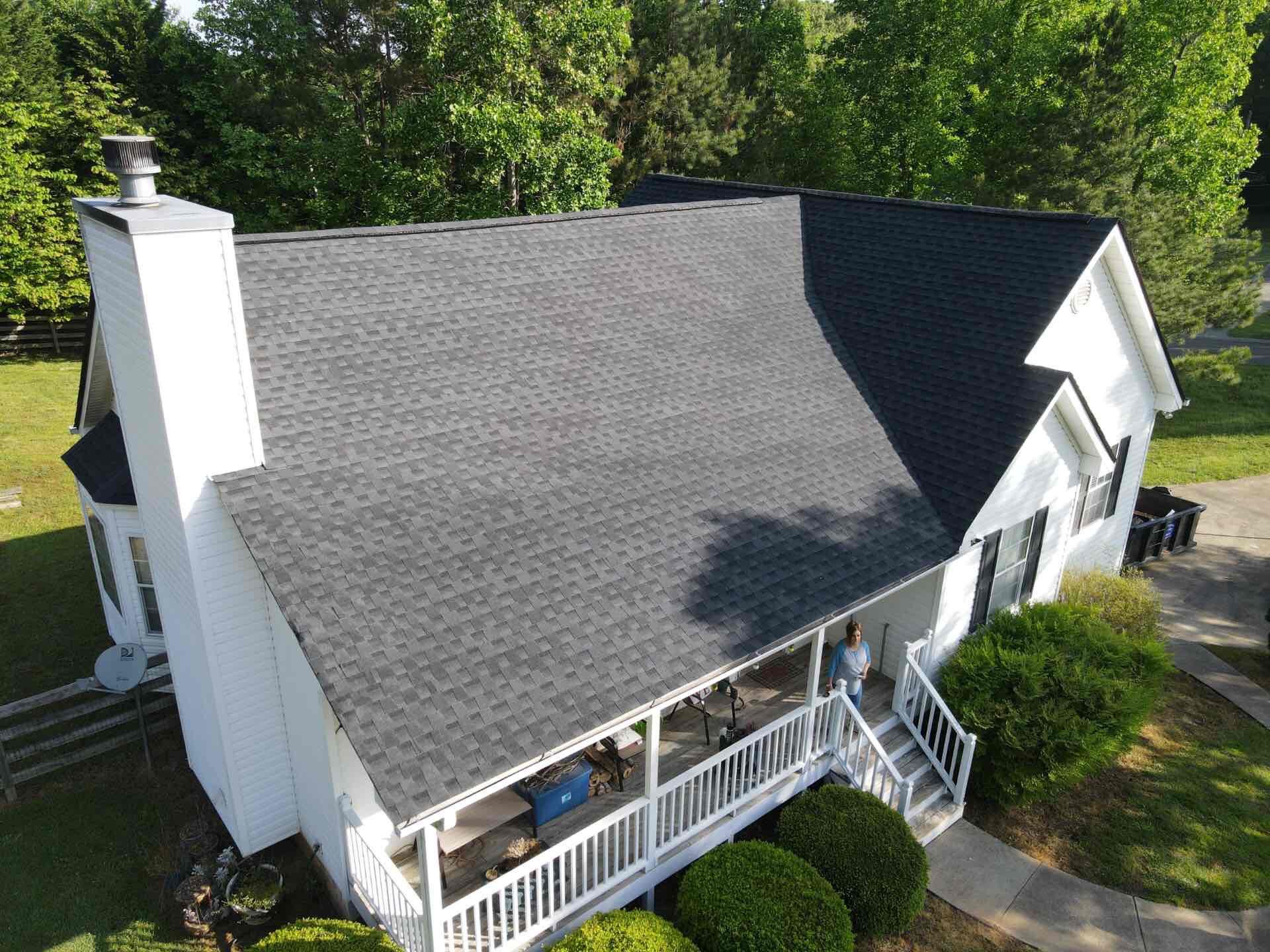 White house with dark gray roof, chimney, and porch, surrounded by greenery.