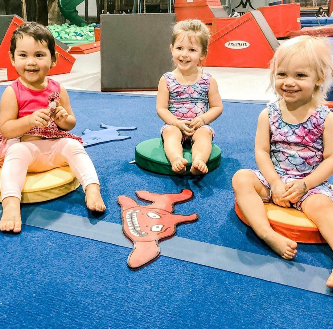 Three young children sit on colorful mats, smiling at the camera in a gym setting.